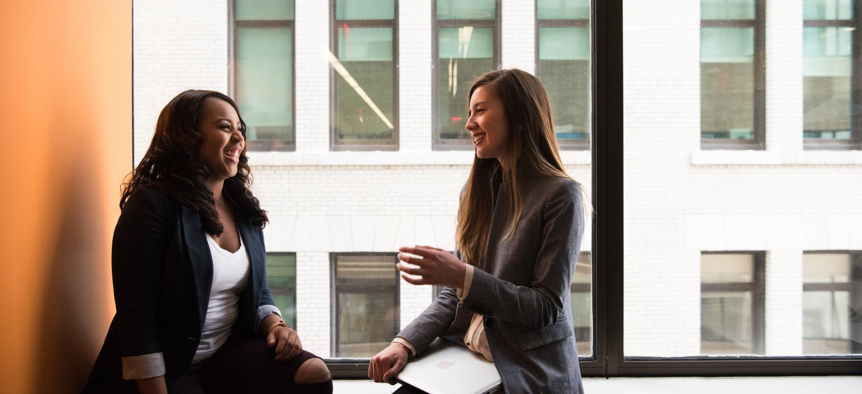 Two young women in blazers sitting and talking, women to the right is sitting with a laptop. Window in the background, looking out onto another building.