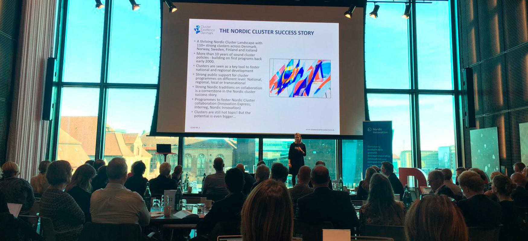 People sitting in front of a stage with a large screen at the Nordic Cluster Meet-up 2018.